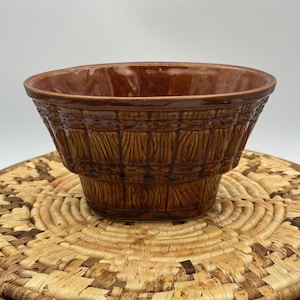 May include: A brown ceramic bowl with a textured, basket-weave design. The bowl has a wide, open top and sits on a woven placemat. The bowl's exterior features vertical and horizontal lines, creating a rustic aesthetic.
