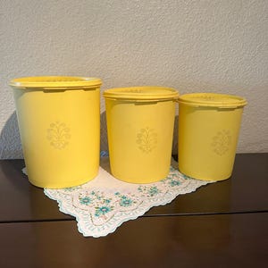 May include: A set of three yellow vintage canisters with lids. Each canister has a decorative floral design. The canisters are different sizes and are arranged on a floral doily on a dark brown surface.