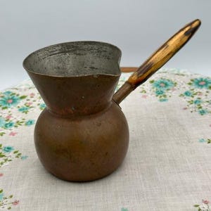 May include: A vintage copper coffee pot with a wooden handle. The pot has a bulbous base and a flared top. The handle is attached to the side of the pot and is made of wood. The pot is likely used for making Turkish coffee.