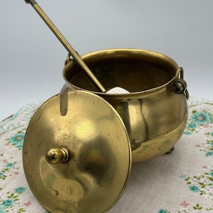 May include: A vintage brass cauldron with a lid and a long-handled spoon. The cauldron has a rounded body, small feet, and a handle on each side. The lid is slightly ajar, revealing the interior. The spoon rests inside the cauldron.