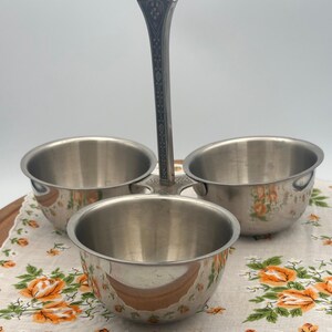 May include: A three-bowl serving dish with a decorative handle. The stainless steel bowls are arranged around a central handle with floral detailing. The bowls sit on a floral patterned cloth with orange roses.