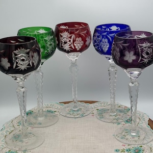 May include: Six colorful wine glasses with intricate floral and geometric cut designs. The glasses feature ruby red, green, blue, and purple bowls, with clear, textured stems and bases. The glasses are arranged on a floral patterned cloth.
