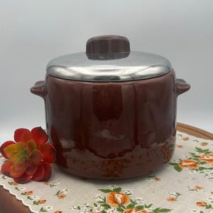 May include: A brown ceramic bean pot with a silver lid and a brown knob. The pot is sitting on a floral patterned fabric.
