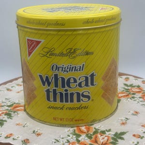 May include: A yellow metal tin of Nabisco Wheat Thins snack crackers. The tin has a diagonal striped pattern and the words "Limited Edition" and "Original Wheat Thins" in large text. The net weight is 13 oz.