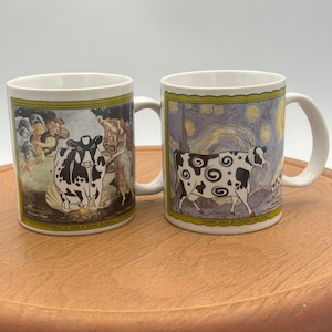 May include: Two white ceramic mugs with cow-themed artwork. One mug features a black and white cow in front of a classical painting, the other a cow with a starry night background. Both mugs have a green border.