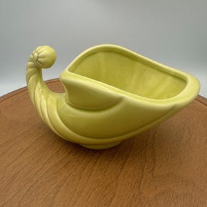 May include: A light green ceramic cornucopia-shaped planter. The planter has a curved, horn-like shape with a rounded end and a wide opening. The surface is smooth and glossy, with a slight variation in color.