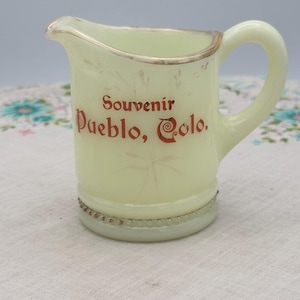 May include: A pale yellow glass creamer pitcher featuring a gold-trimmed rim and a beaded base. The front displays the red text Souvenir Pueblo, Colo. with faint, light-colored floral accents etched into the surface of the glass.