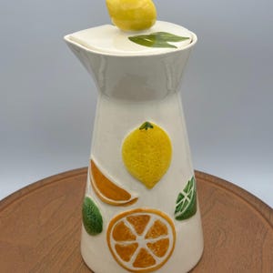 May include: A white ceramic pitcher with a lid, adorned with embossed citrus fruit designs. The lid features a yellow lemon handle. The pitcher has a tall, slender shape and is decorated with orange and green fruit slices.