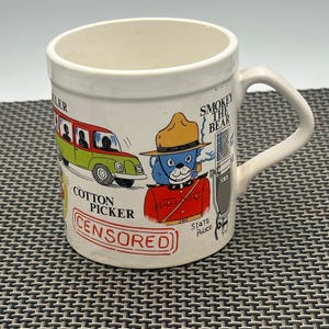 May include: White ceramic mug with a cartoon illustration of Smokey the Bear, a cotton picker, and a state police officer. The mug has the text "Smokey the Bear", "Cotton Picker", "State Police", and "CENSORED".