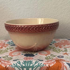 May include: A cream-colored ceramic mixing bowl with a maroon heart-shaped design around the rim. The bowl has a flared shape and a decorative design on the front. The bowl is sitting on a patterned tablecloth with coral and sea life.