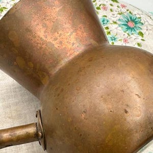 May include: A vintage copper pitcher with a long handle. The pitcher has a wide base that tapers to a narrower neck and a flared rim. The copper has a warm, aged patina with areas of darker oxidation.