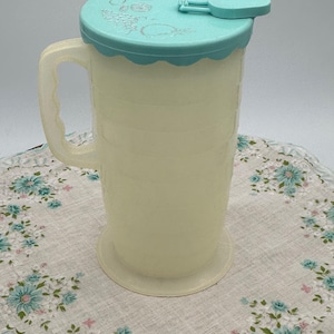 May include: A vintage, off-white plastic pitcher with a light blue lid and handle. The lid has a scalloped edge and floral detailing. The pitcher has measurement markings and sits on a round base. The handle is curved and attached to the side.