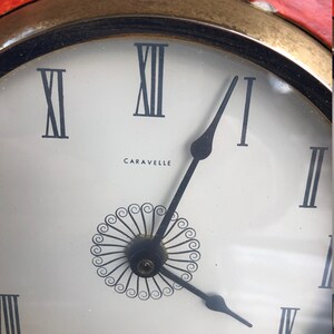 Puede incluir: Una esfera de reloj blanca con marcadores de hora romanos negros y agujas negras. La esfera del reloj tiene un dise&ntilde;o decorativo negro en el centro y la palabra "Caravelle" impresa debajo.