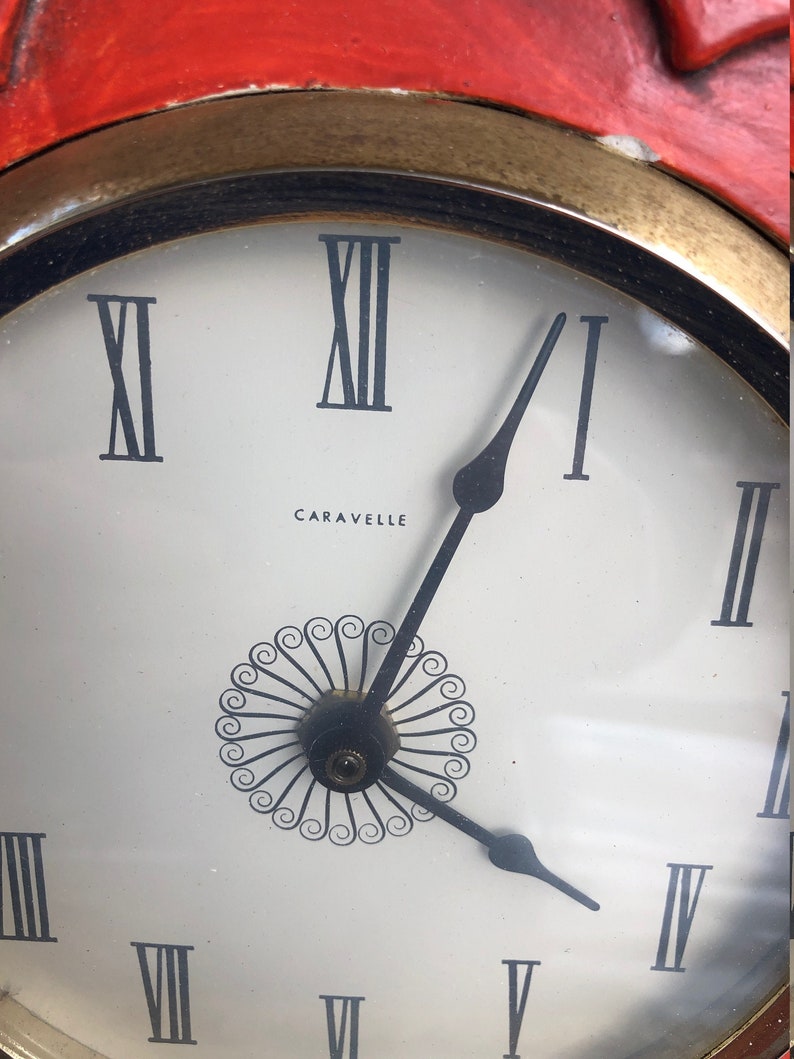 Puede incluir: Una esfera de reloj blanca con marcadores de hora romanos negros y agujas negras. La esfera del reloj tiene un dise&ntilde;o decorativo negro en el centro y la palabra "Caravelle" impresa debajo.