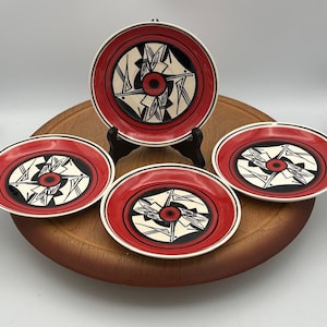 May include: Set of four decorative ceramic plates with a red border and a central design in black, white, and red. One plate is displayed on a black stand, while the others are arranged on a wooden surface.
