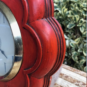 Puede incluir: Un reloj de madera rojo con un borde dorado y una esfera blanca. El reloj tiene forma de flor con cinco p&eacute;talos.