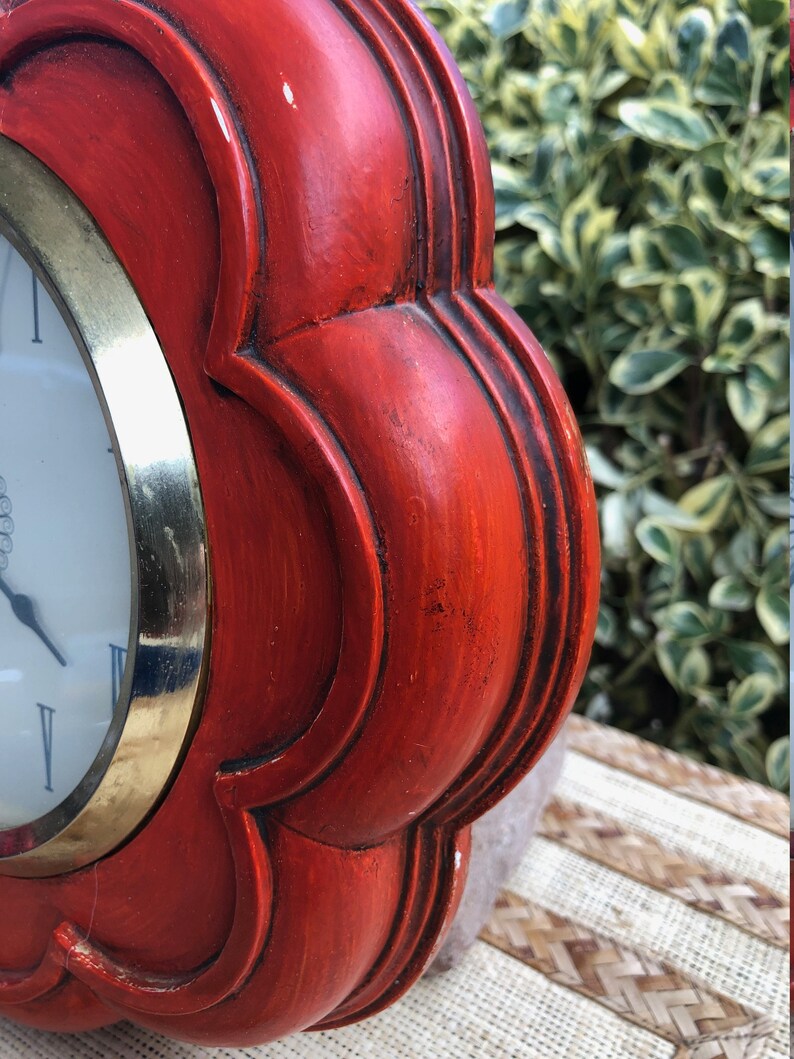 Puede incluir: Un reloj de madera rojo con un borde dorado y una esfera blanca. El reloj tiene forma de flor con cinco p&eacute;talos.