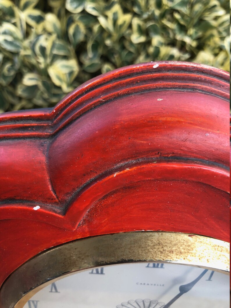 Puede incluir: Un reloj de madera rojo con un borde dorado y una esfera blanca. La esfera del reloj tiene n&uacute;meros romanos y la palabra "Caravelle" impresa en ella.