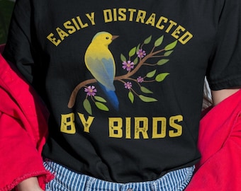 May include: Black t-shirt with the text "Easily Distracted By Birds" in yellow letters. A yellow bird with blue wings is perched on a branch with pink flowers.