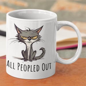 May include: White ceramic mug featuring a grumpy cartoon cat with yellow eyes and the text "ALL PEOPLED OUT" in black. The cat is gray and seated. The mug has a curved handle.
