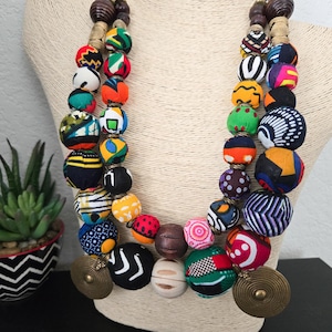 May include: A colourful, multi-strand necklace with large, fabric-covered beads in various patterns and hues. The necklace incorporates wooden beads and brass-coloured circular pendants. The necklace is displayed on a mannequin.