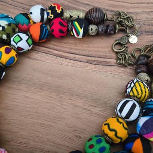 May include: A vibrant necklace featuring fabric-covered beads in diverse patterns and colours, such as red, blue, yellow, and black. The necklace also includes wooden and metal beads, and a bronze-coloured chain.