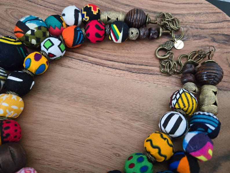 May include: A vibrant necklace featuring fabric-covered beads in diverse patterns and colours, such as red, blue, yellow, and black. The necklace also includes wooden and metal beads, and a bronze-coloured chain.