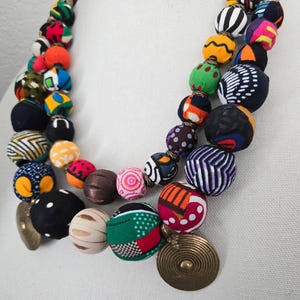 May include: A vibrant necklace featuring large, fabric-covered beads in a variety of patterns and colours. The beads are arranged in two strands and are complemented by gold-toned, circular pendants. The necklace showcases a mix of geometric and floral designs.