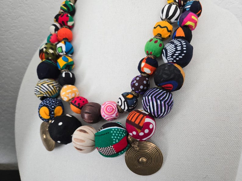 May include: A vibrant necklace featuring large, fabric-covered beads in a variety of patterns and colours. The beads are arranged in two strands and are complemented by gold-toned, circular pendants. The necklace showcases a mix of geometric and floral designs.