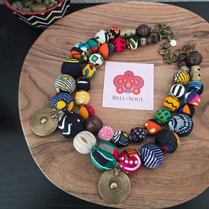 May include: A vibrant beaded necklace with large, patterned beads in various colours and designs. The necklace features a brass pendant and clasp, and a small card with the text "BELL+SOUL". The necklace is displayed on a wooden plate.