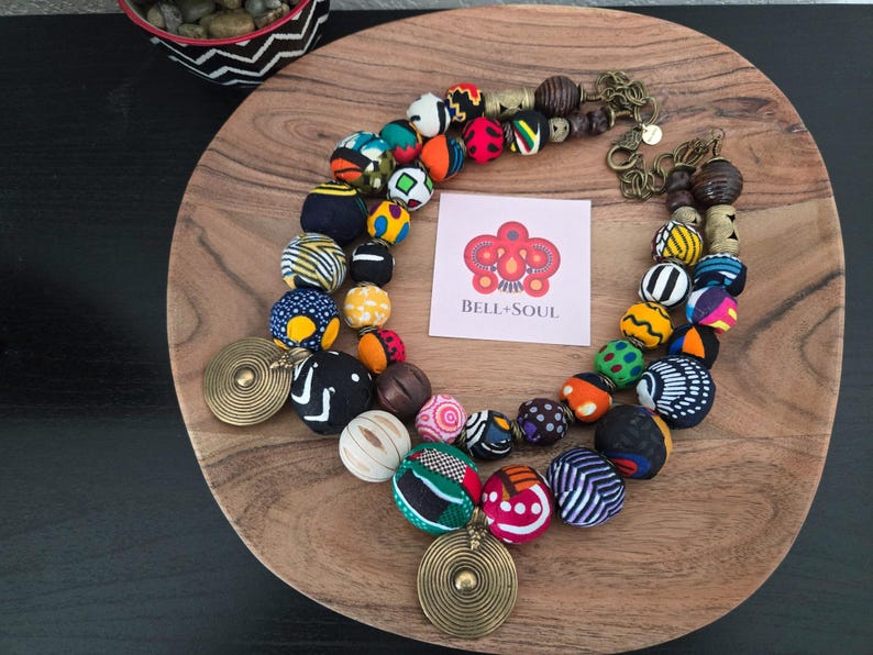 May include: A vibrant beaded necklace with large, patterned beads in various colours and designs. The necklace features a brass pendant and clasp, and a small card with the text "BELL+SOUL". The necklace is displayed on a wooden plate.