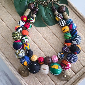 May include: A vibrant beaded necklace featuring large, patterned beads in a variety of colours, including red, blue, yellow, and black. The necklace includes wooden beads and brass-coloured circular pendants. The beads are arranged in a double strand.