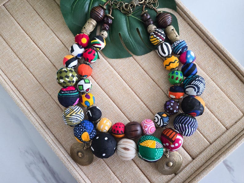 May include: A vibrant beaded necklace featuring large, patterned beads in a variety of colours, including red, blue, yellow, and black. The necklace includes wooden beads and brass-coloured circular pendants. The beads are arranged in a double strand.
