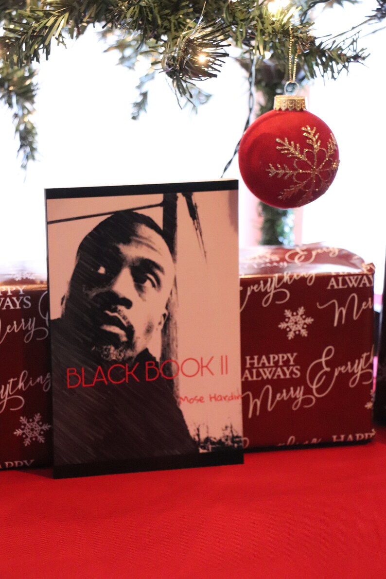 K&ouml;nnte beinhalten: Ein Schwarzwei&szlig;foto eines Mannes auf dem Cover eines Buches mit dem Titel "Black Book II" von Mose Hardin. Das Buch liegt auf einer roten Oberfl&auml;che mit einem roten Weihnachtsbaumschmuck und einer verpackten Geschenkbox mit einer wei&szlig;en Schneeflocke und den Worten "Happy Always Merry Everything" in wei&szlig;er Kursivschrift.