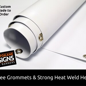 May include: White vinyl banner with grommets and a heat-welded hem. The banner is rolled up and shows the grommets and hem. The text "Custom Made to Order" and "SPOKANE SIGNS & BANNERS" is visible on the banner.
