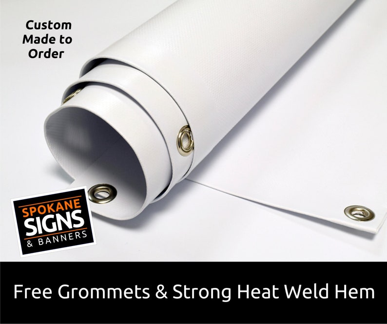 May include: White vinyl banner with grommets and a heat-welded hem. The banner is rolled up and shows the grommets and hem. The text "Custom Made to Order" and "SPOKANE SIGNS & BANNERS" is visible on the banner.