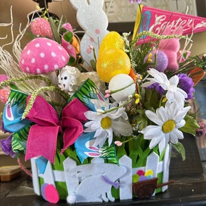 Puede incluir: Una cesta de Pascua de valla blanca llena de flores, un conejito blanco, un hongo rosa con lunares blancos y huevos de Pascua coloridos. La cesta está decorada con un lazo rosa y una cinta azul.