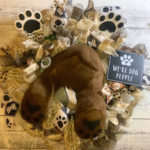 May include: A brown dog-shaped wreath with a black and white paw print design. The wreath is decorated with brown, beige, and black mesh and ribbons. A black sign with white text reads "We're Dog People".