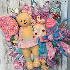 May include: A gingerbread person doll wearing a pink and white sweater and skirt, holding a pink and white candy cane lollipop. The doll is surrounded by pink, white, and blue ribbon, candy canes, and other holiday decorations. The sign says "Baked with love".