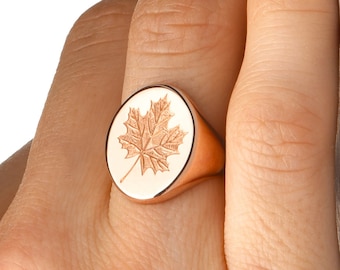 May include: Rose gold signet ring with a detailed maple leaf design. The ring has a polished finish and an oval shape. The maple leaf is engraved on a cream-coloured background, creating a contrast with the rose gold.