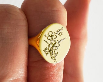 May include: A gold signet ring featuring an engraved floral design. The ring is worn on a finger with red nail polish.