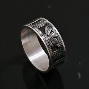 Peut inclure: Une bague en argent avec un motif gravé d'aigle et de motifs géométriques. La bague a une large bande et une finition polie, mettant en valeur des détails complexes. La bague est un bijou.