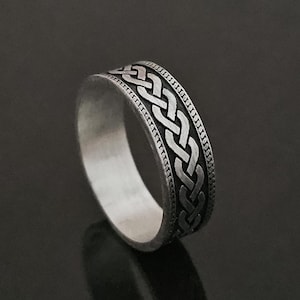 Puede incluir: Anillo de plata con diseño de nudo celta. El anillo presenta un patrón trenzado detallado alrededor de la banda. El anillo se muestra sobre una superficie negra reflectante.