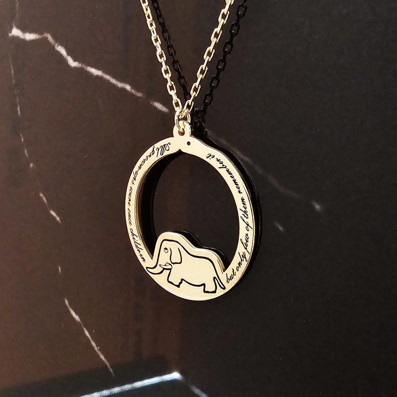 K&ouml;nnte beinhalten: Goldkette mit einem runden Anh&auml;nger, der eine Elefanten-Silhouette und die Inschrift "Erwachsene waren einst Kinder, aber nur wenige erinnern sich daran" zeigt.