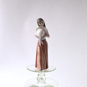 Könnte beinhalten: Eine Porzellanfigur einer Frau in einem braunen Kleid, die einen weißen Hut mit einem rosa Band hält.