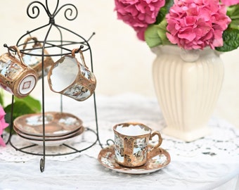 May include: A set of four delicate porcelain teacups and saucers with gold accents and floral patterns. The cups are displayed on a black metal stand and a saucer is placed on a white lace tablecloth. A bouquet of pink hydrangeas in a white vase is in the background.