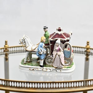 以下が含まれることがあります： 3体のフィギュアが乗った馬車を描いた磁器製の置物。馬車は赤と金で、屋根は白。フィギュアは18世紀の服装をしている。馬は白で金色のアクセントが施されている。