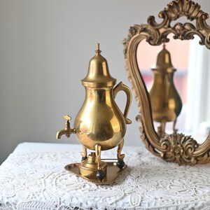 Puede incluir: Una tetera de latón dorado con un pico y una manija, sentada sobre un soporte decorativo con tres patas. La tetera está sobre un mantel de encaje blanco.
