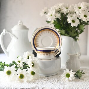 Könnte beinhalten: Eine weiße Teetasse und Untertasse mit goldenen und blauen Verzierungen, eine weiße Teekanne und ein weißer Milchkännchen auf einer weißen Spitzentischdecke mit weißen Gänseblümchen.
