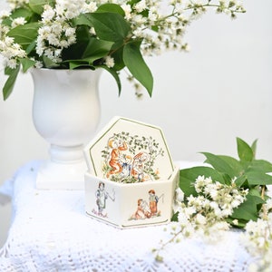 Może przedstawiać: Białe, sześciokątne pudełko ceramiczne z pokrywką. Pudełko jest ozdobione zielonym i białym wzorem kwiatowym oraz sceną dzieci bawiących się w ogrodzie. Pudełko stoi na białym haftowanym obrusie.